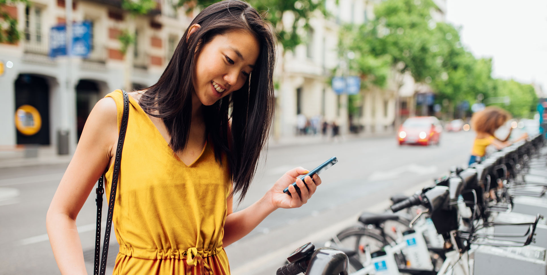 Eine lächelnde junge Frau hält ein Handy und steht vor einer Reihe von Rädern eines Bike-Sharing-Anbieters.
