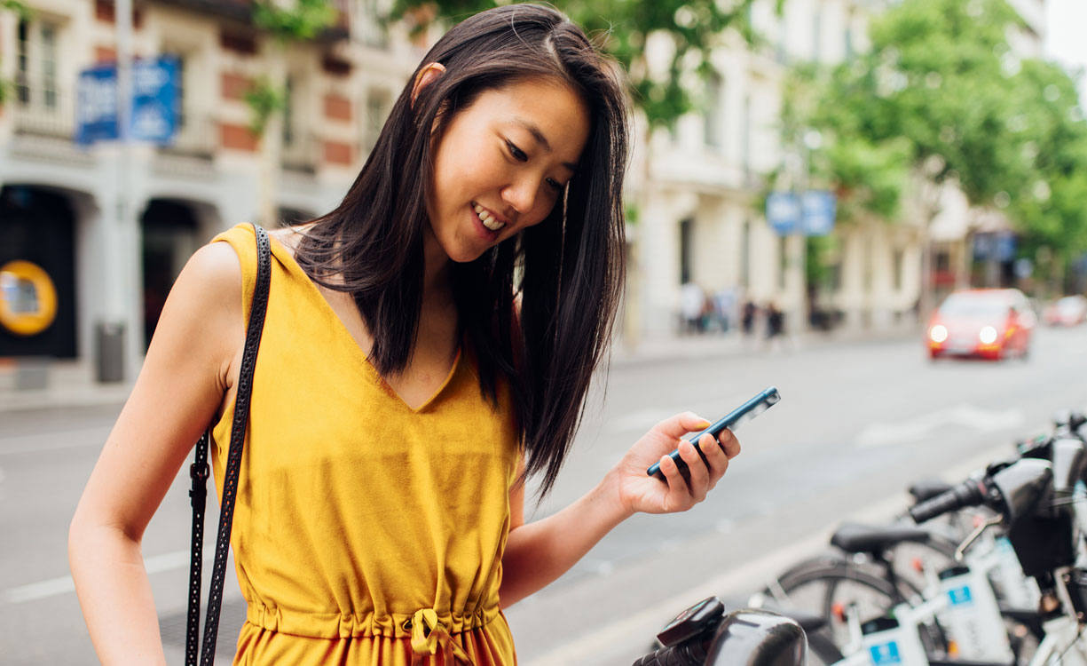 Eine lächelnde junge Frau hält ein Handy und steht vor einer Reihe von Rädern eines Bike-Sharing-Anbieters.