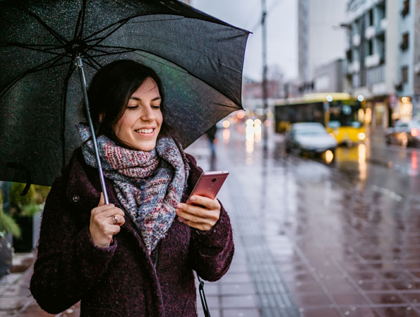 Junge Frau unter Regenschrim sieht sich den Bildschirm ihres Handys an.