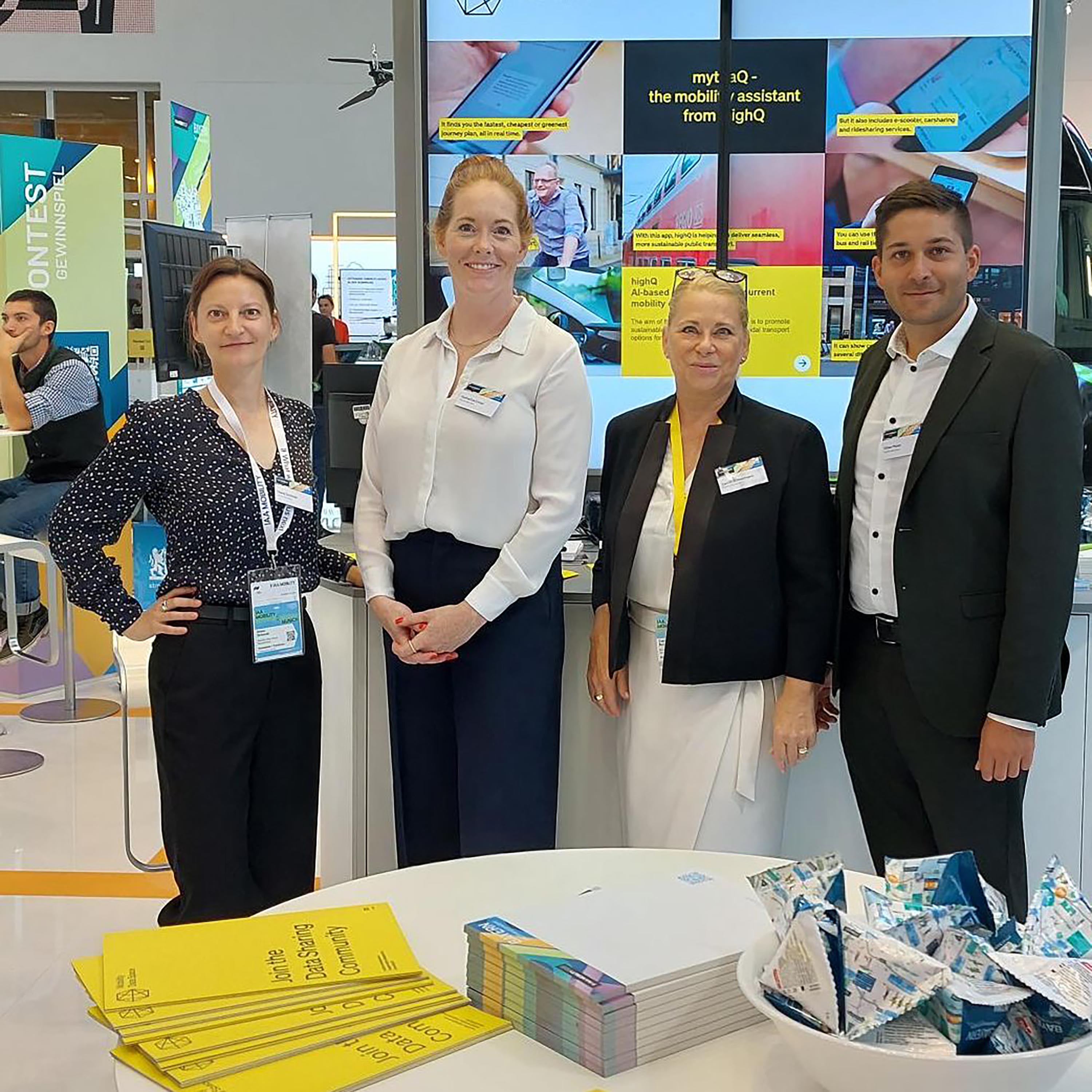 The picture shows four people standing next to each other, smiling at the camera. They are at a trade fair stand. In the background is a screen showing a presentation. In the foreground there is a table with flyers and small promotional gifts.