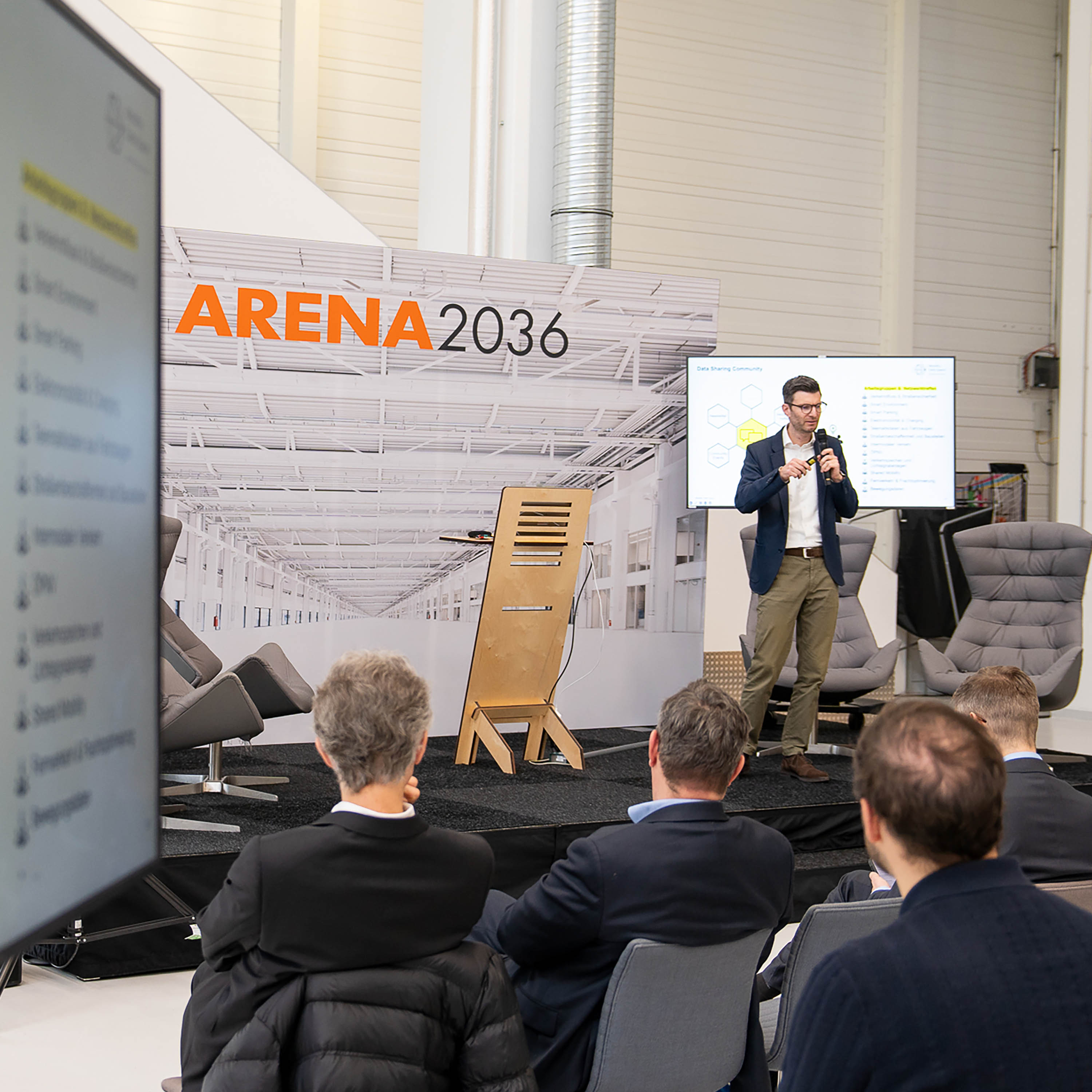 The picture shows a small stage on which a man is speaking into a microphone. Behind him there is a banner saying "ARENA 2036" and a television showing a presentation. The audience is seated on chairs in front of him. On the far left of the picture is a second screen showing the same presentation as on the stage.