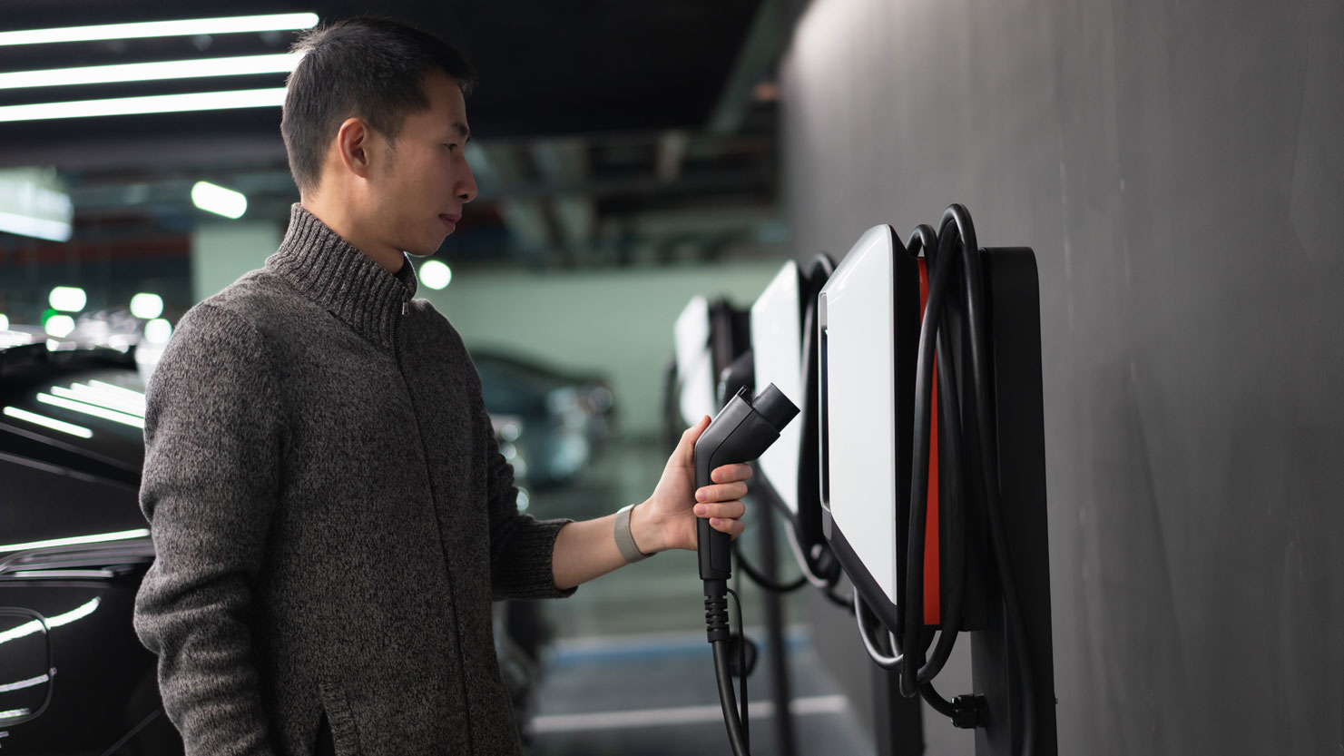 Man standing at a charging station for electric vehicles in a parking garage, holding a charging plug in his left hand. In the background, several cars parked in a row.
