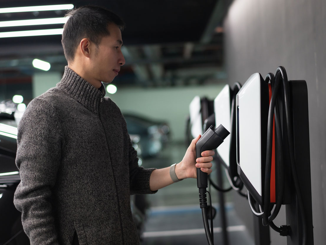 Man standing at a charging station for electric vehicles in a parking garage, holding a charging plug in his left hand. In the background, several cars parked in a row.