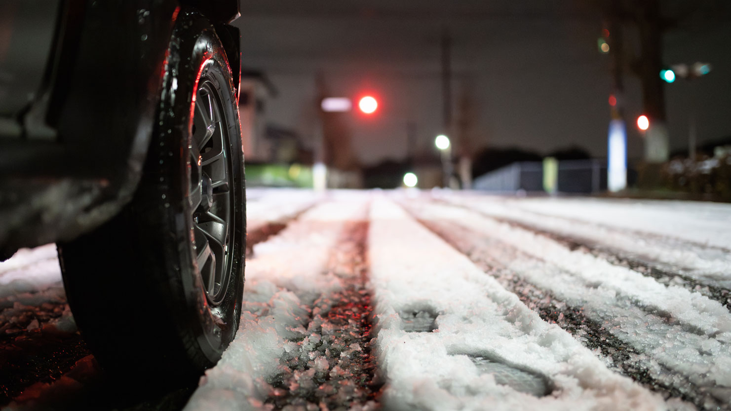 Close-up of the tyre of a car on a snow-covered road. Dusky surroundings, occasional lights visible in the background.