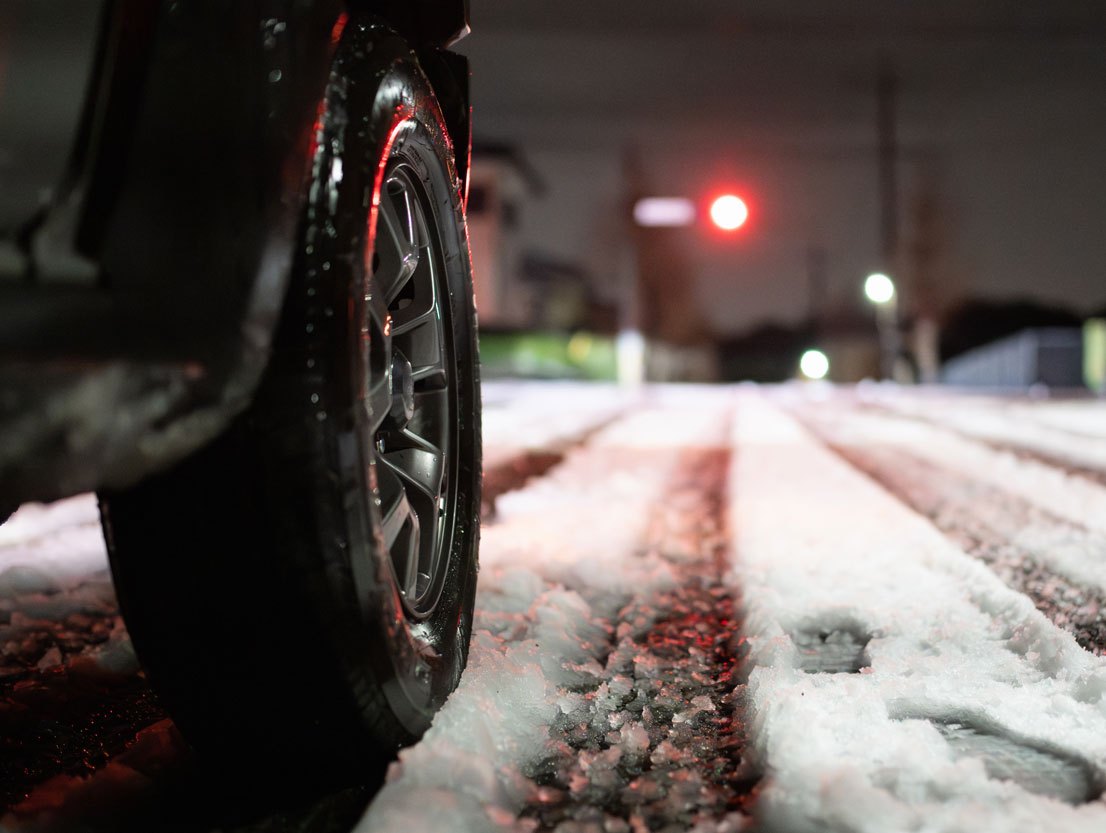 Close-up of the tyre of a car on a snow-covered road. Dusky surroundings, occasional lights visible in the background.