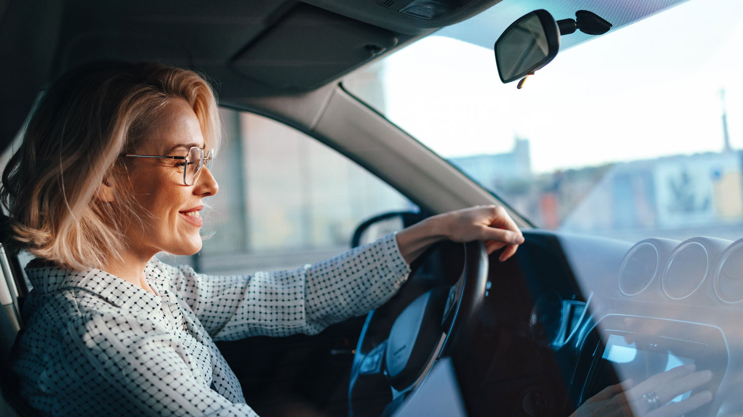 Frau sitzt lächelnd auf dem Fahrersitz eines Autos und hat die linke Hand auf das Lenkrad gelegt. Aufnahme aus der Perspektive des Beifahrers. 
