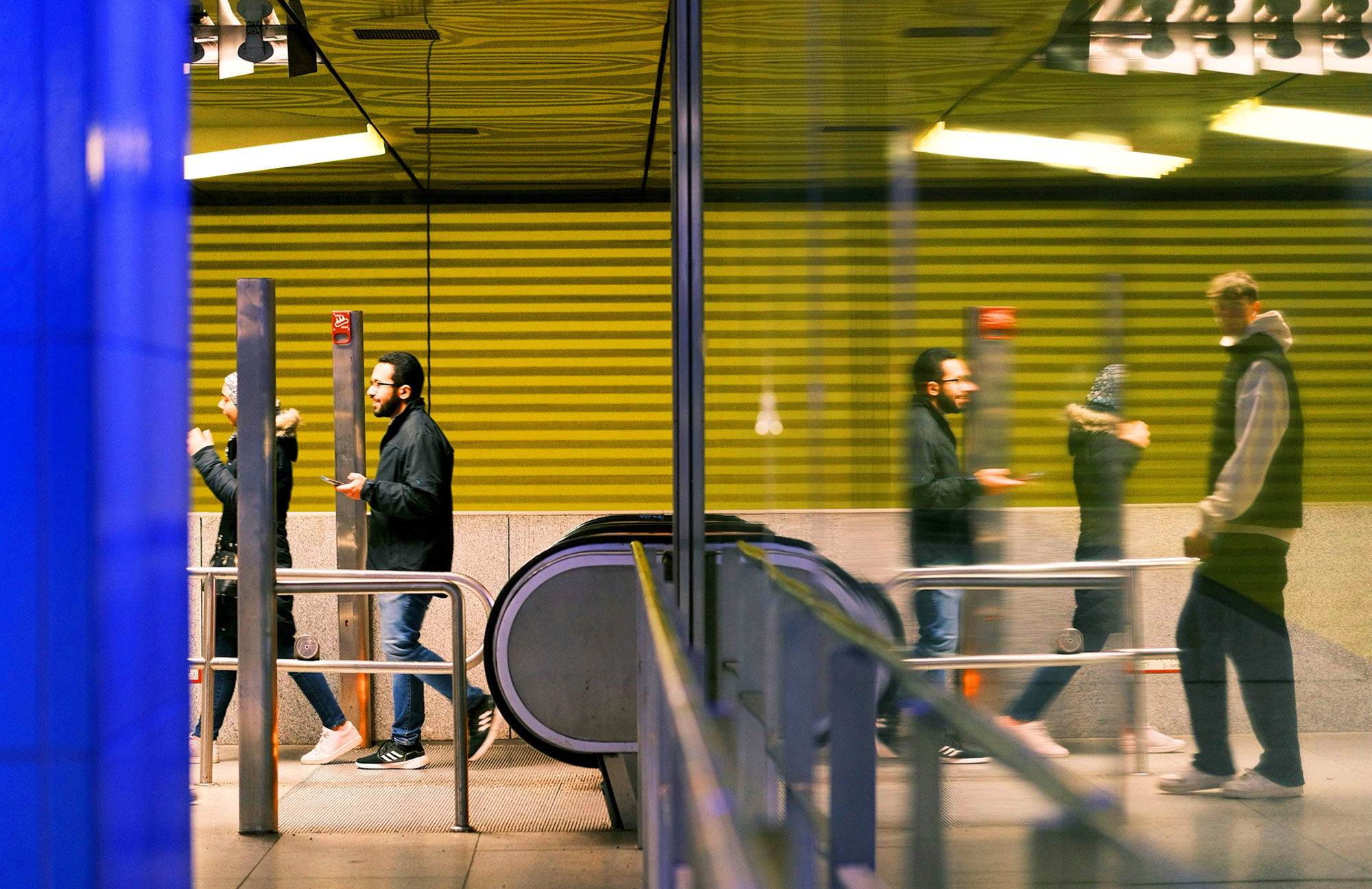 The picture shows a cross-section of a railway staircase with an escalator. On the left are two people who have just arrived at the top, one of them holding a mobile phone. On the right is a glass reflection in which another person can be seen.